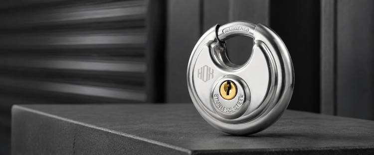 Silver HOX keyed disc padlock on a charcoal ledge in a softly lit storage setting.
